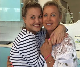 Two women hugging and smiling in a kitchen, one wearing a striped shirt, the other a floral lace top.