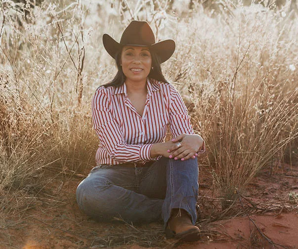 A woman in a striped shirt and cowboy hat sitting in a grassy field, smiling.