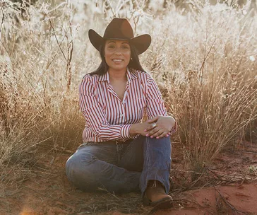 A woman in a striped shirt and cowboy hat sitting in a grassy field, smiling.