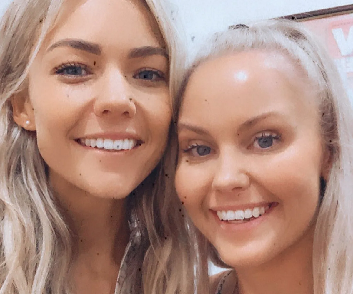 Two smiling women with long, light-colored hair take a close-up selfie indoors.