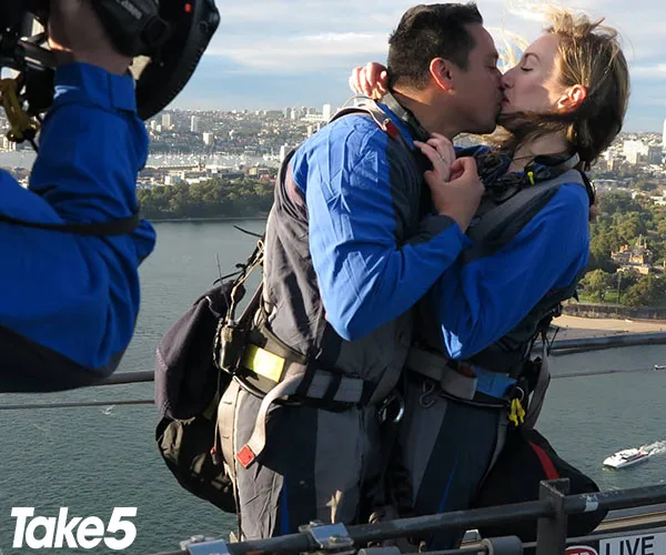 Couple kissing on Sydney Harbour Bridge, wearing safety gear, with cityscape and water visible in the background.
