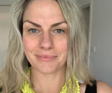 A woman with long blond hair wearing a bright yellow beaded necklace, smiling softly at the camera.