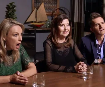 Three people sit at a table reacting with surprise and interest, a wooden ship model seen in the background.