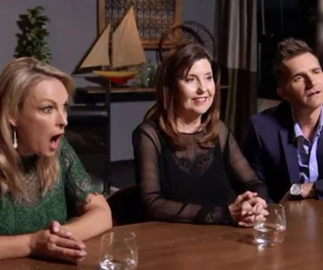 Three people sit at a table reacting with surprise and interest, a wooden ship model seen in the background.