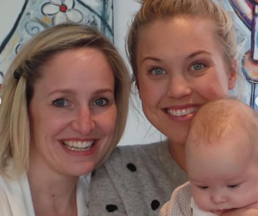 Two smiling women with a baby in their arms, in front of a colorful wall art background.