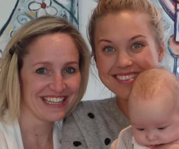 Two smiling women with a baby in their arms, in front of a colorful wall art background.