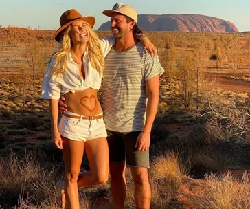 A couple smiling in a desert landscape; woman showing a heart drawn on her midriff.