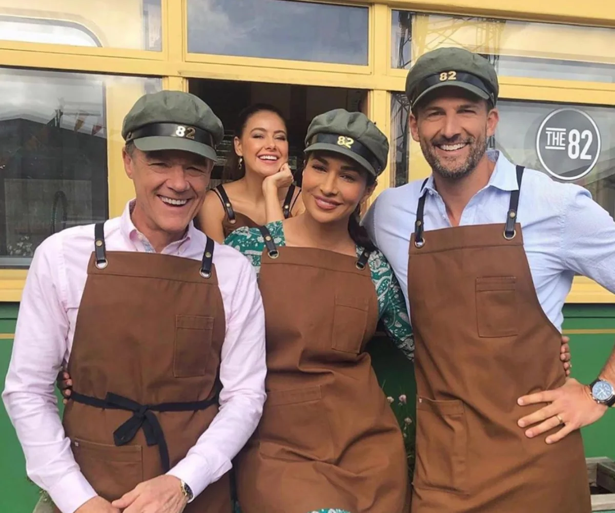 Four people in brown aprons and green hats pose happily outside a retro cafe with a sign reading "The 82".
