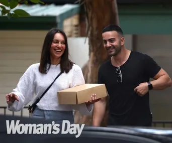A smiling woman and man carrying a box walking outdoors with a "Woman's Day" logo at the bottom.
