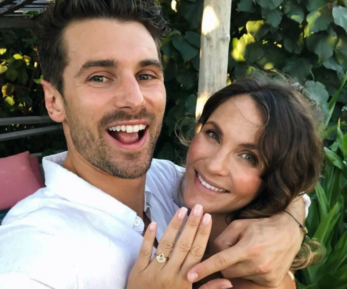 A smiling couple poses outdoors, with the woman showing off an engagement ring.