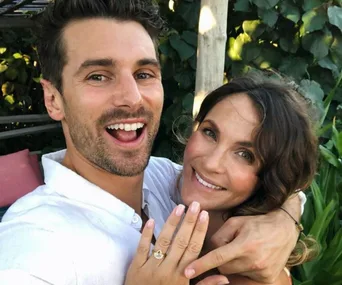 A smiling couple poses outdoors, with the woman showing off an engagement ring.