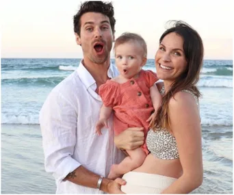 Family smiling at the beach, with the father holding a toddler and the mother showing a baby bump.