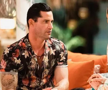 Man in floral shirt sitting on a couch talking, with tropical decor in the background.