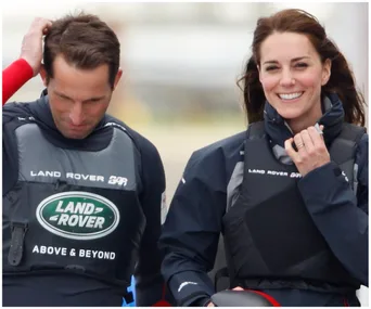 Kate Middleton and a man in Land Rover sailing gear, smiling on a windy day during a sports event.