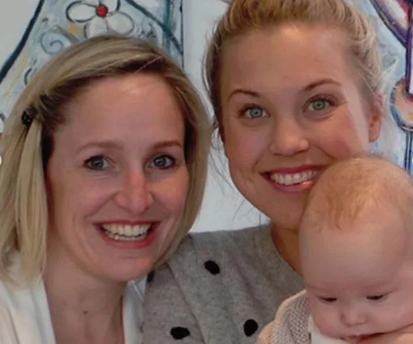 Two smiling women pose closely with a baby in front, indoors with artwork in the background.