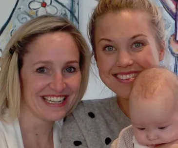 Two smiling women pose closely with a baby in front, indoors with artwork in the background.