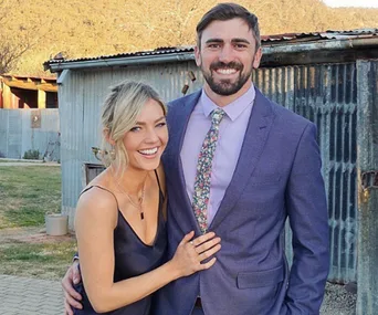 A happy couple, the woman in a black dress and the man in a suit, stand together smiling outside a rustic building.