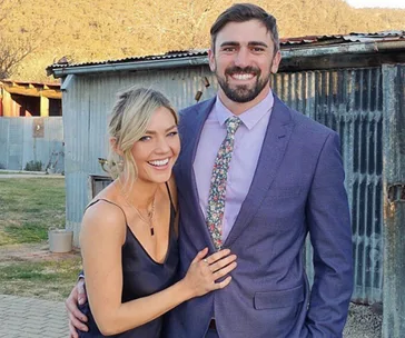 A happy couple, the woman in a black dress and the man in a suit, stand together smiling outside a rustic building.