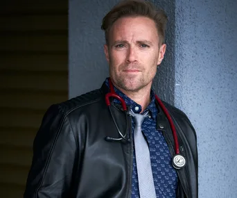 Man in a leather jacket with a stethoscope around his neck, standing against a textured blue wall.