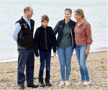Family of four smiling on a pebble beach, with the sea in the background, dressed casually in outdoor wear.