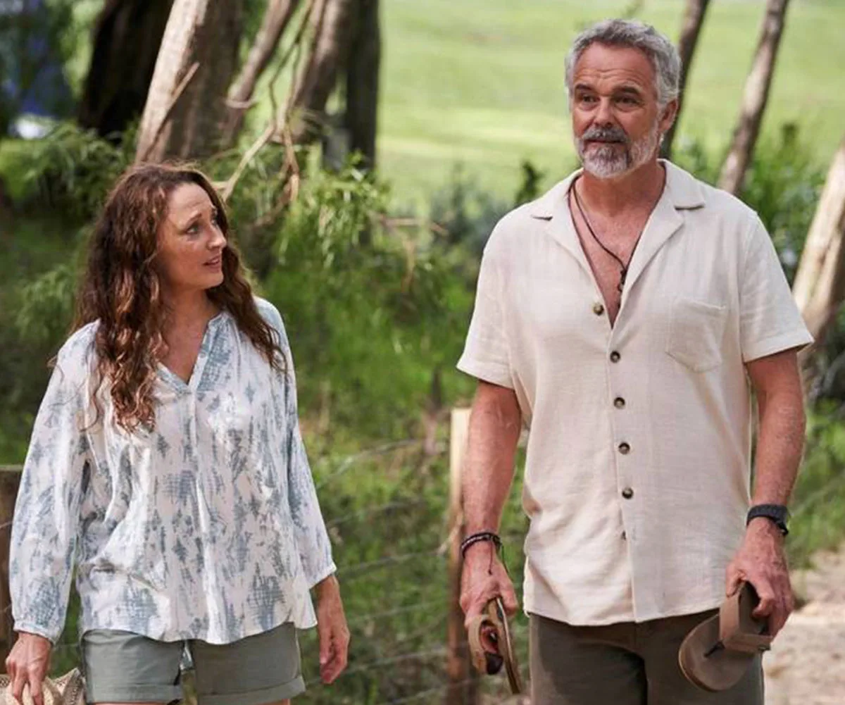 A man and a woman walk through a leafy, wooded area, both in casual summer attire, carrying sandals.