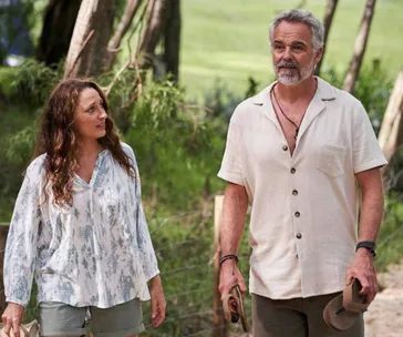 A man and a woman walk through a leafy, wooded area, both in casual summer attire, carrying sandals.