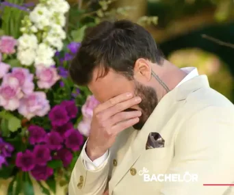 Man in a beige suit covers his face with hand, standing in front of colorful flowers, emotional moment from "The Bachelor."
