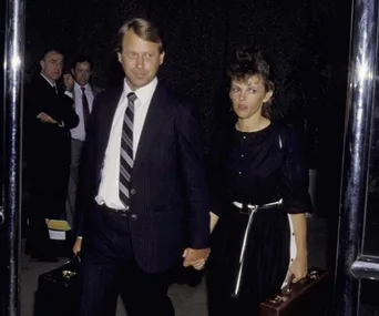 A couple in formal attire, holding hands, exits a building; man in suit with briefcase, woman in black dress with white belt.