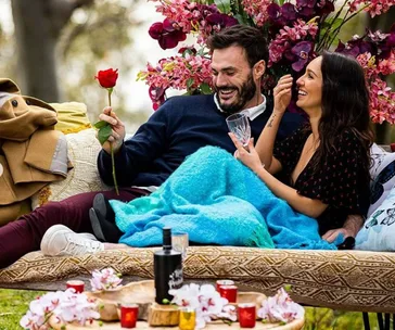 Man holding a rose and smiling with a woman on a couch outdoors, surrounded by pink flowers and drinks.