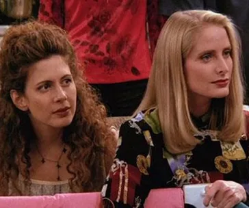 Two women sitting, one with curly hair and the other with straight, both wearing patterned outfits, looking forward.