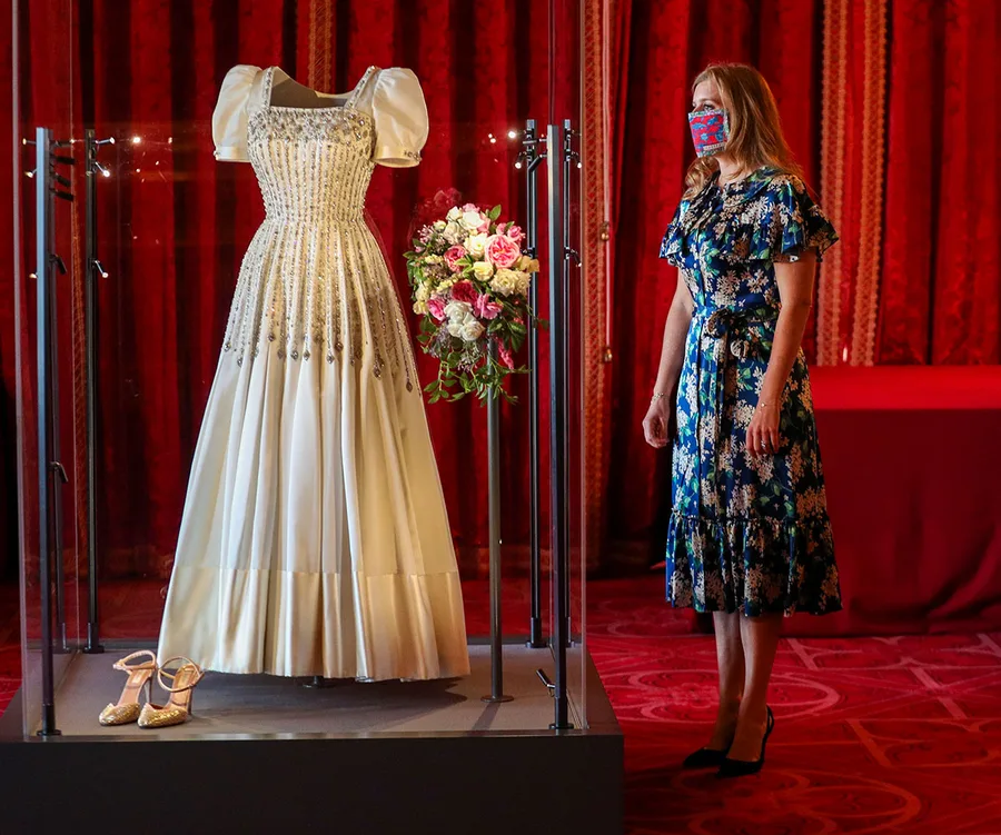 Princess Beatrice's wedding dress display with floral backdrop; a woman in a floral dress observes closely.