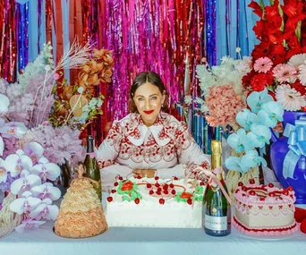 A woman in a patterned dress behind a table with decorative cakes, flowers, and champagne, against a colorful backdrop.