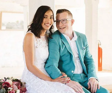 A smiling couple in formal attire, seated together. The woman wears a white dress, and the man wears a teal suit.