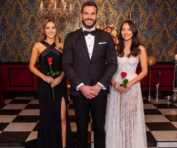 Three people standing in formal attire, two women holding roses, in an ornately decorated room with a chandelier.