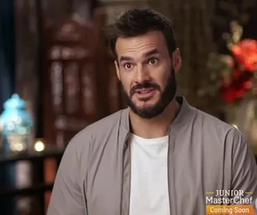 Man with a beard and light shirt speaks in an indoor setting, with a Junior MasterChef logo on screen.