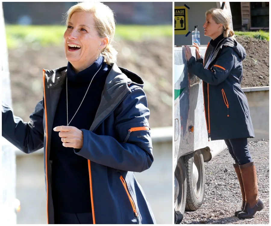 Woman in outdoor jacket and boots interacting cheerfully at a farm, standing by a trailer, and holding its side.