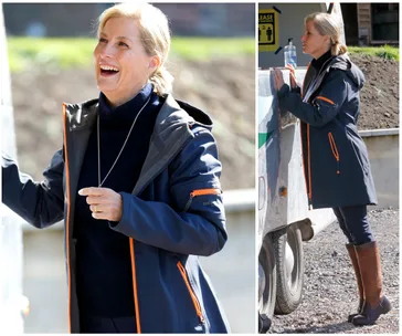 Woman in outdoor jacket and boots interacting cheerfully at a farm, standing by a trailer, and holding its side.