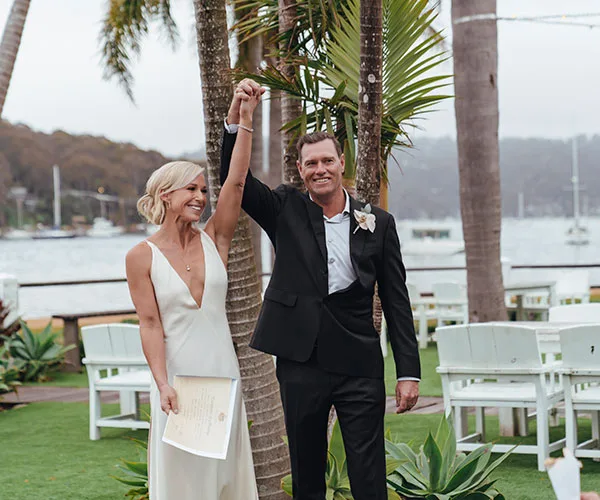 Wedding couple celebrates under palm trees by a waterfront with chairs and greenery in the background.
