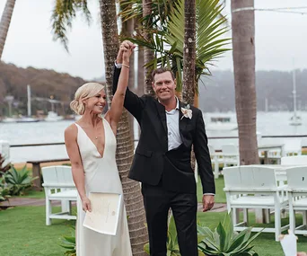 Wedding couple celebrates under palm trees by a waterfront with chairs and greenery in the background.
