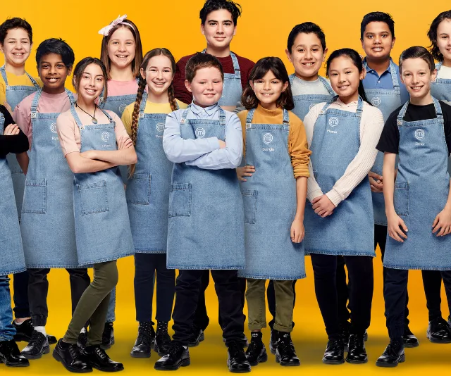 Group of Junior MasterChef 2020 contestants wearing blue aprons, smiling in front of a yellow background.