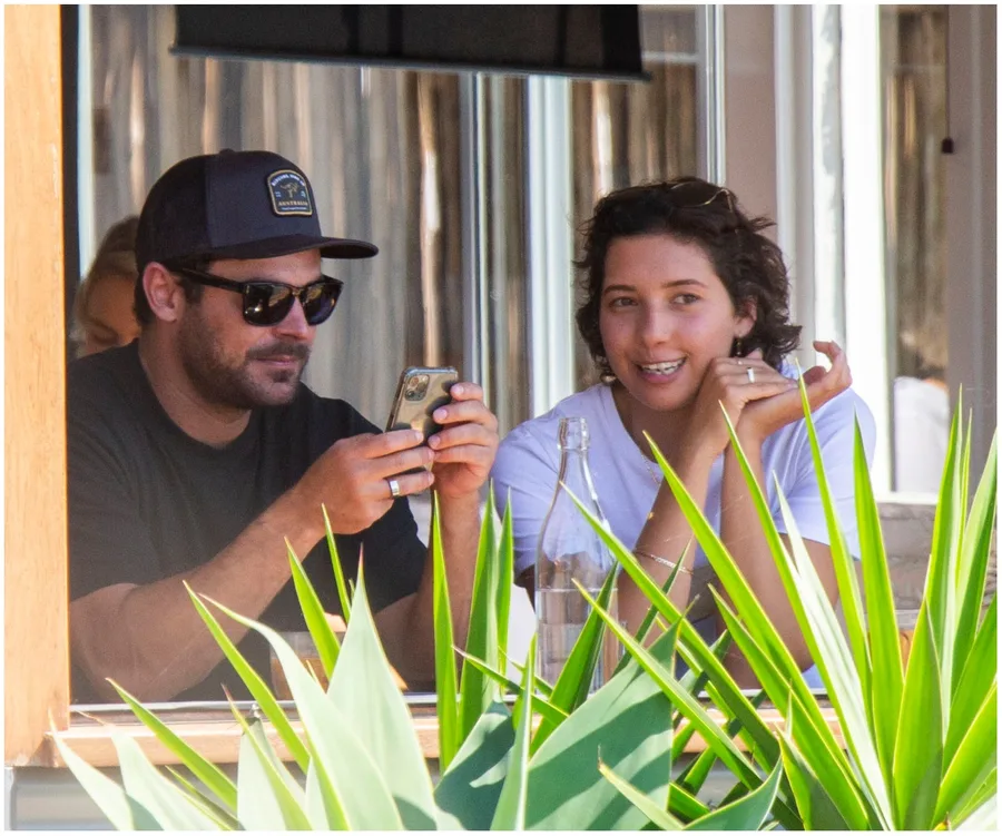 Two people sitting at an outdoor café, one holding a phone, with plants in the foreground.