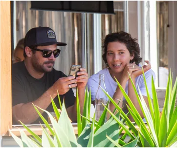 Two people sitting at an outdoor café, one holding a phone, with plants in the foreground.