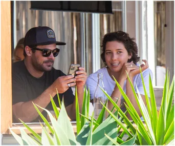 Two people sitting at an outdoor café, one holding a phone, with plants in the foreground.