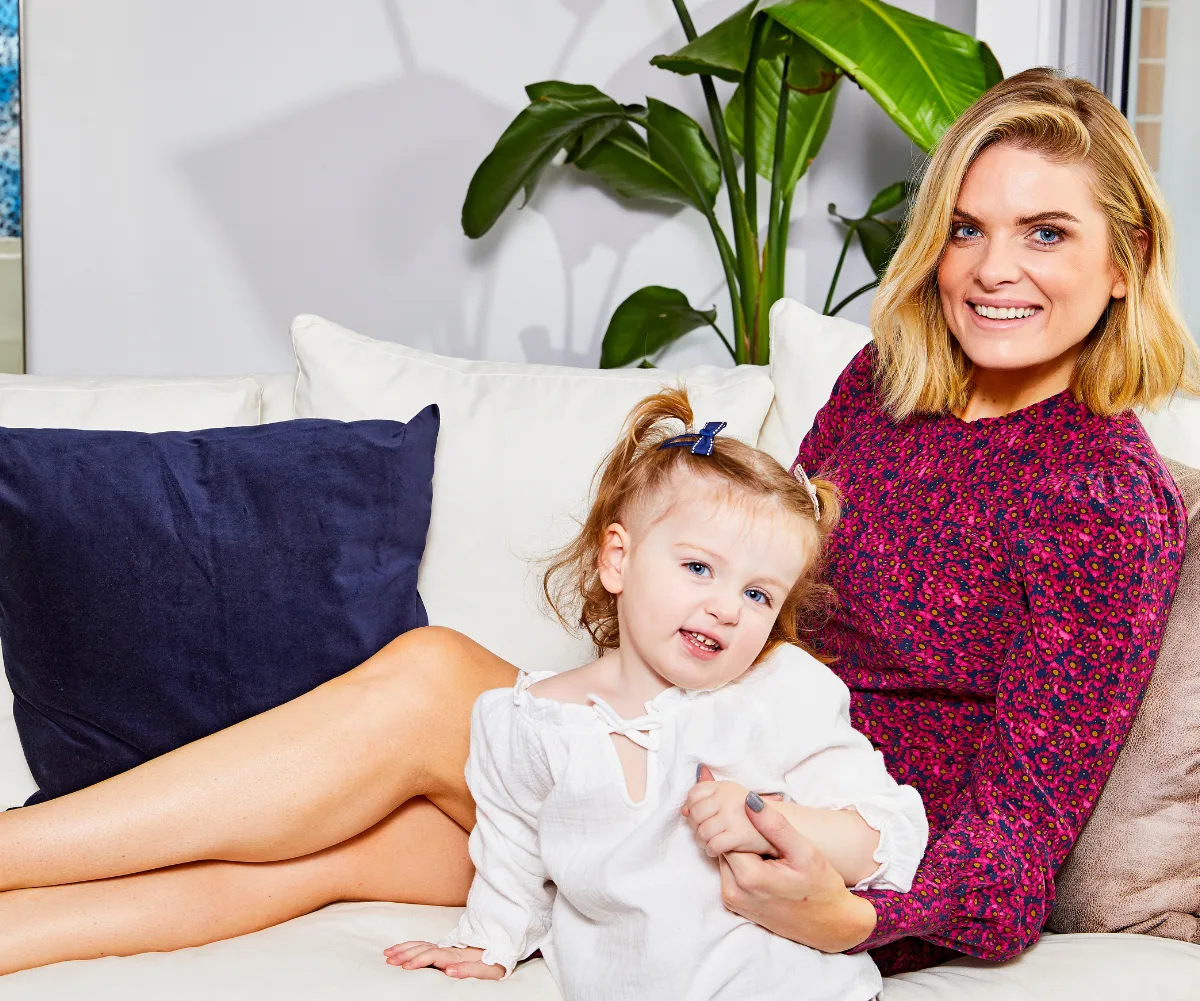 Woman in a red floral dress sitting on a couch with a smiling child in a white top, against a vibrant home backdrop.