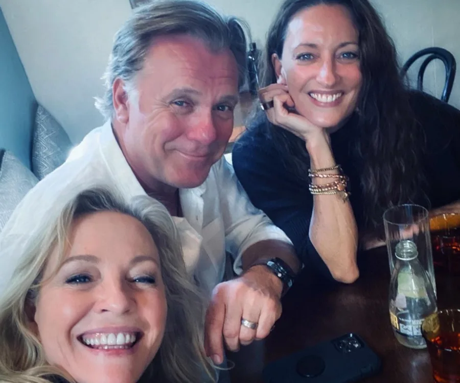 Three people sitting at a table, smiling for a selfie, with drinks and a phone visible on the table.