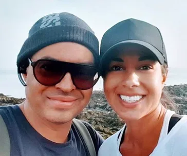 Two people smiling outdoors, wearing hats and sunglasses, with a rocky landscape in the background.