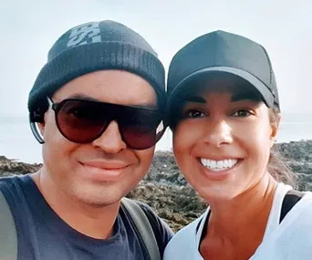 Two people smiling outdoors, wearing hats and sunglasses, with a rocky landscape in the background.