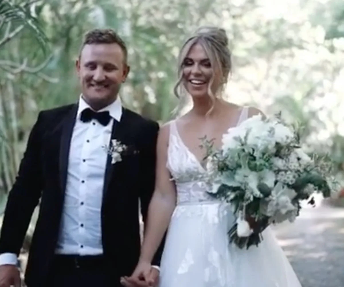 Bride and groom smiling, walking hand-in-hand through a lush, green setting. Bride holds a large bouquet.