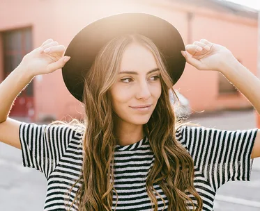 A woman with long, wavy hair wearing a striped shirt and hat smiles outdoors in sunlight.