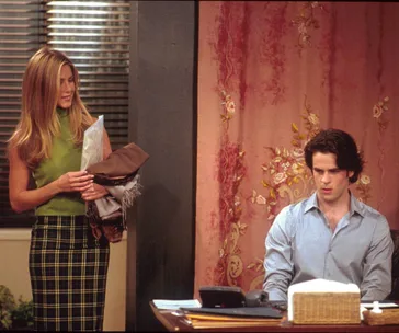 Rachel stands with Gavin, holding clothes, in an office with floral curtains in "Friends."
